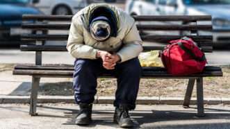 A sad looking man sits outside on a bench hunched over with his elbows on his knees | Photo 141163957 © Srdjan Randjelovic | Dreamstime.com