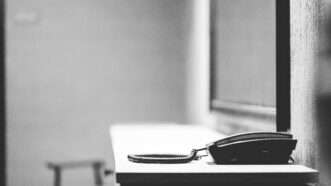 Greyscale photo of a desk phone sitting on a metal table in front of a plexiglass window in a jail. | Wirestock | Dreamstime.com