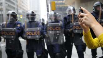 A woman takes pictures on her phone against the backdrop of police in riot gear holding shields. | Arindam Banerjee | Dreamstime.com