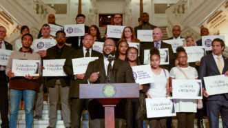 Man gives a speech in front of people holding signs that say Education Freedom | Josh Robertson