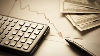 A keyboard and money are seen next to a graph trending downward | Photo 7997381 © Maksym Filipchuk | Dreamstime.com